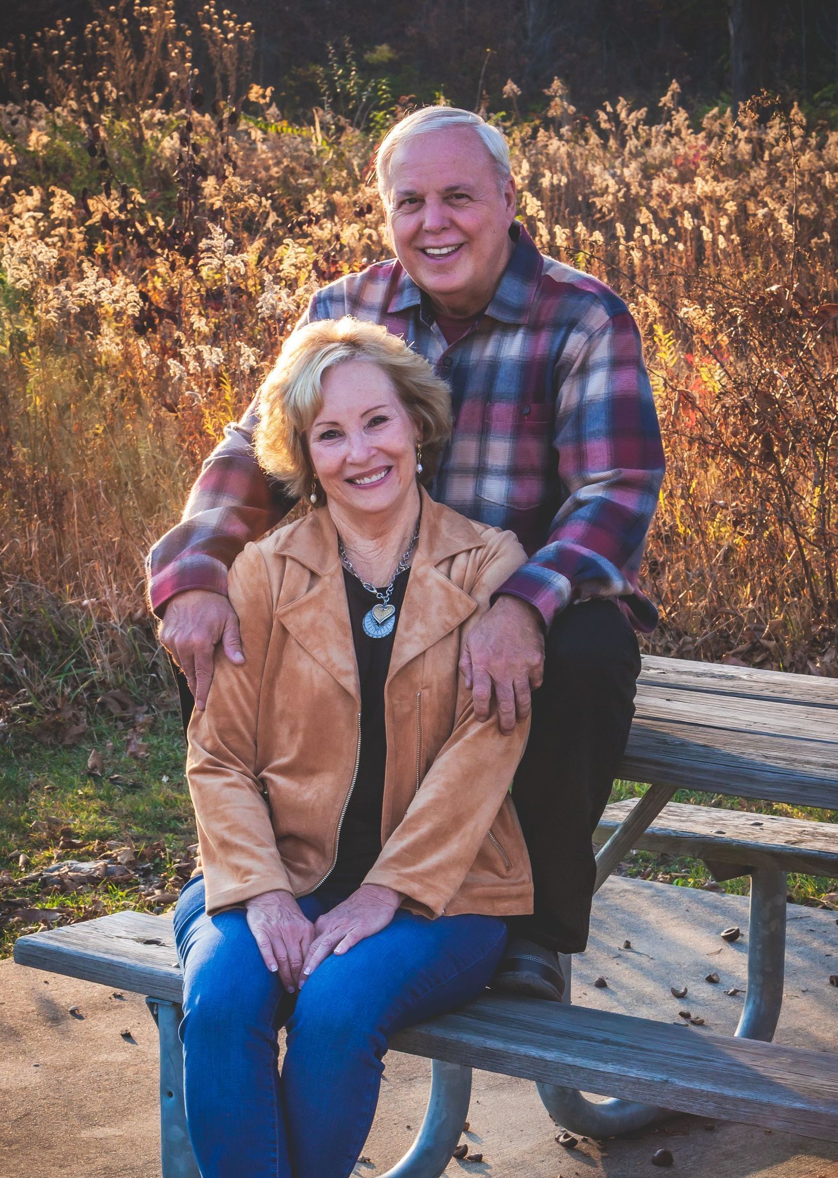 Couple poses on a park bench. Woman in tan jacket, blue jeans smiles; man in plaid shirt smiles behind her.