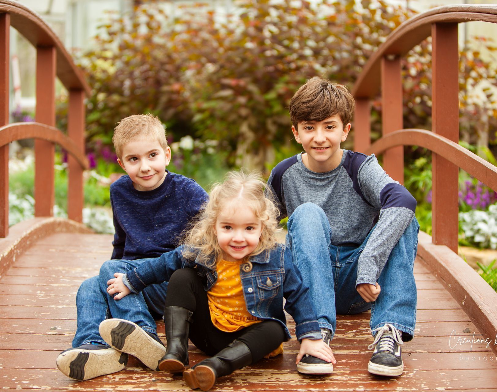 Three children sitting on a wooden bridge; two boys and a girl. Smiling, with a blurred natural background.