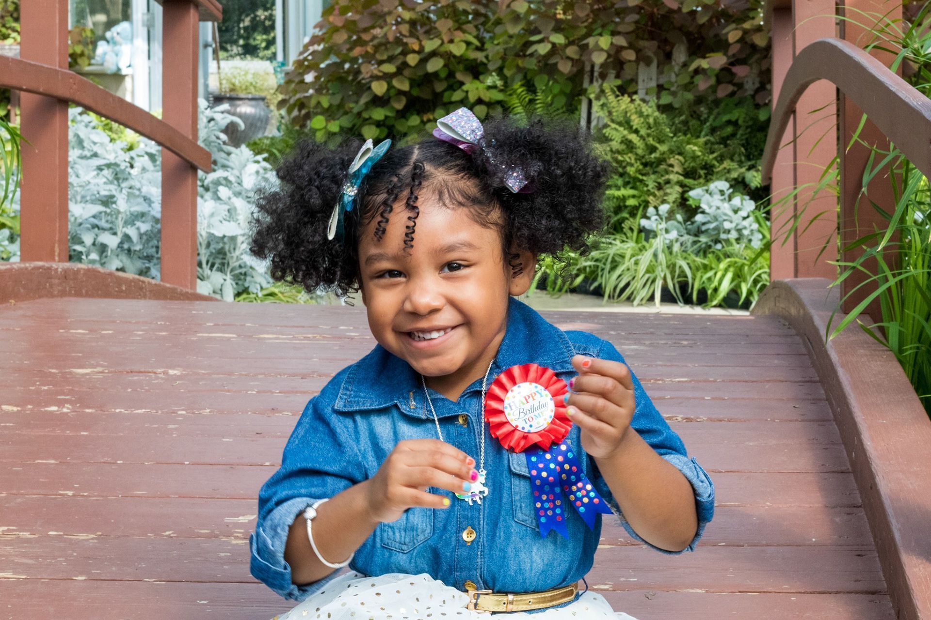 Girl with curly hair, denim shirt, and ribbon smiles on a wooden bridge, outdoors.