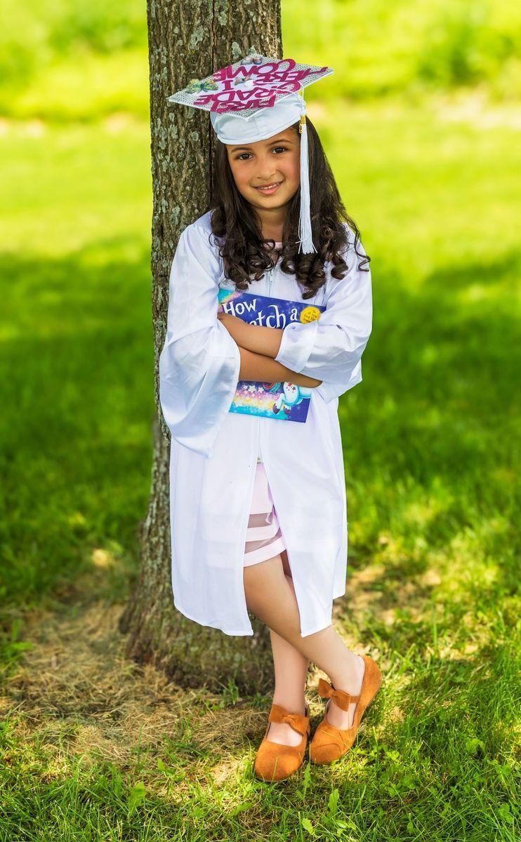 Young person in a white graduation gown and cap, standing near a tree, smiling.