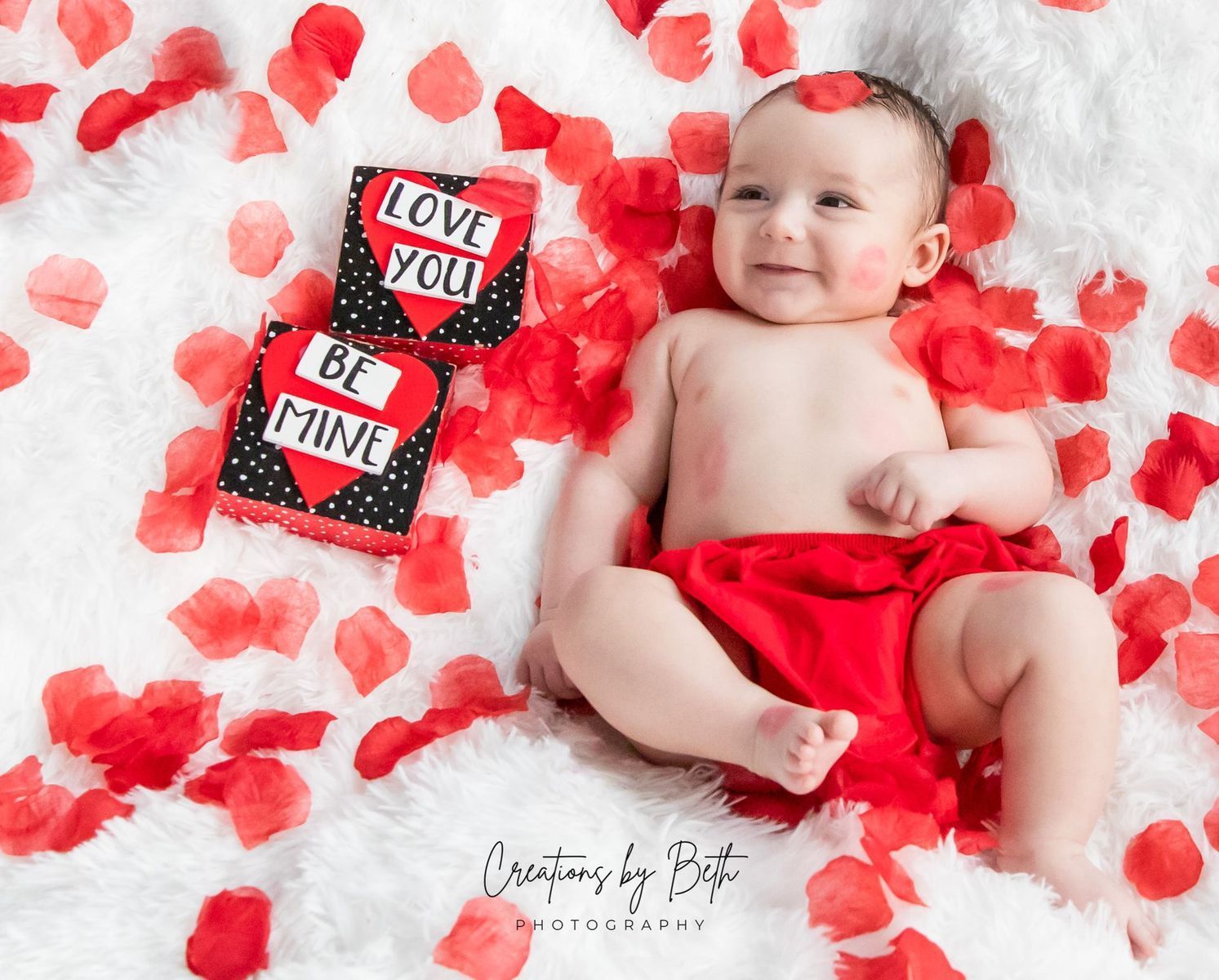 Baby in red diaper and cheek paint, surrounded by rose petals, beside heart-shaped boxes saying 