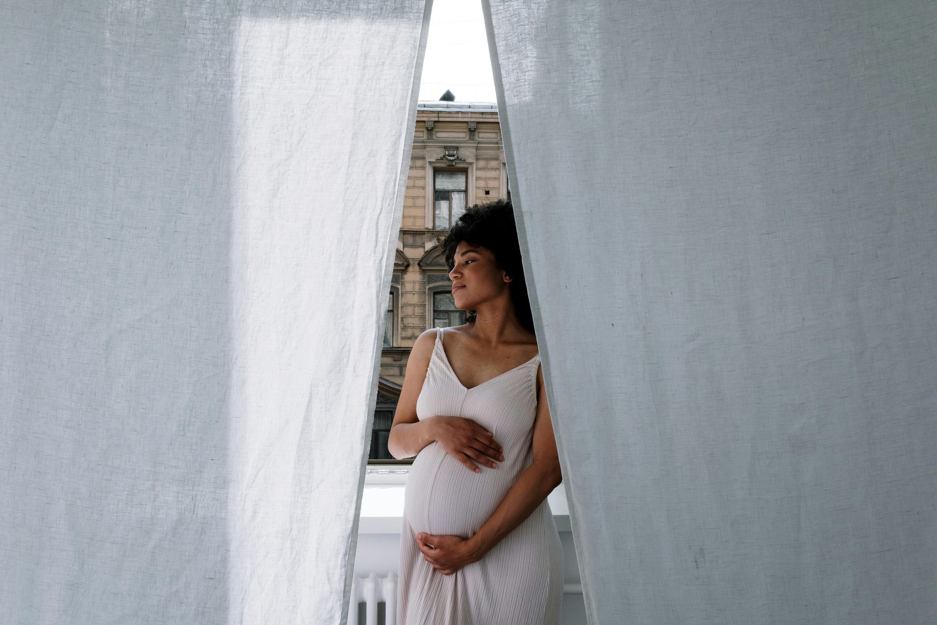 Pregnant person standing between white curtains, wearing a white dress, looking to the side.