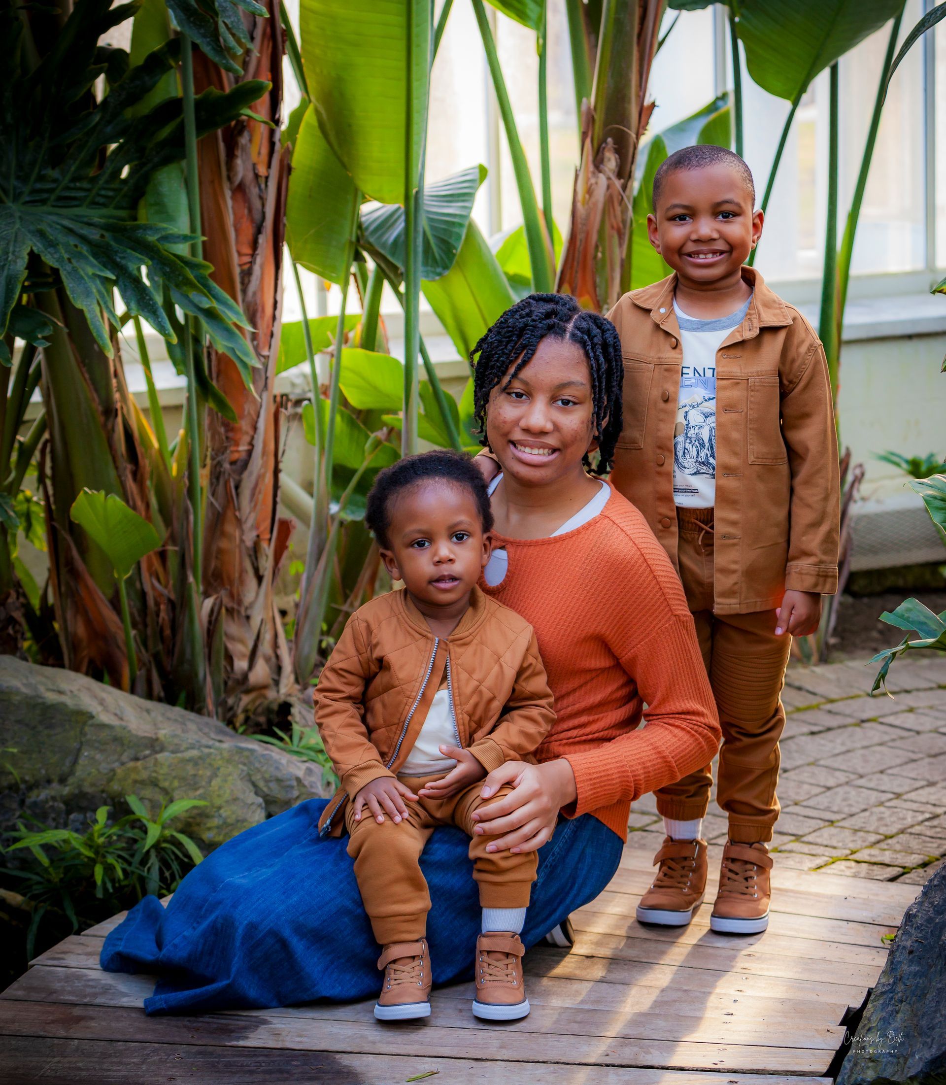 Woman and two children pose by plants. Woman wears orange sweater and blue skirt. Boys wear brown outfits.
