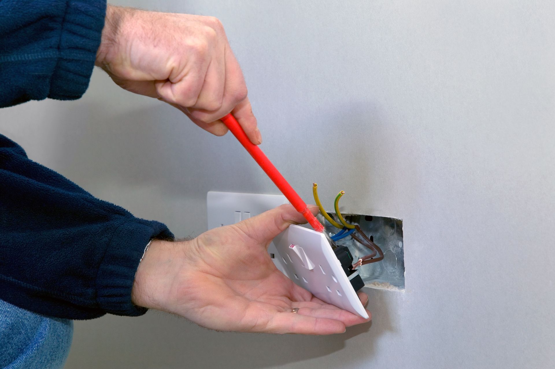 Person removing a white electrical outlet cover with a red screwdriver, wall behind.