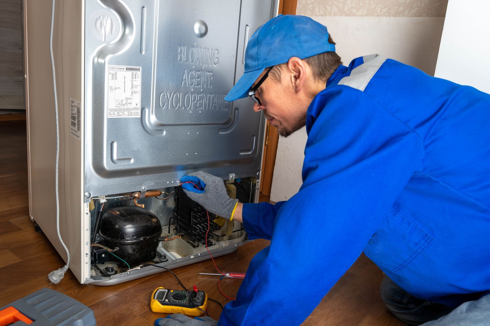 A person in blue overalls repairs a refrigerator's back panel. They wear a cap and gloves.