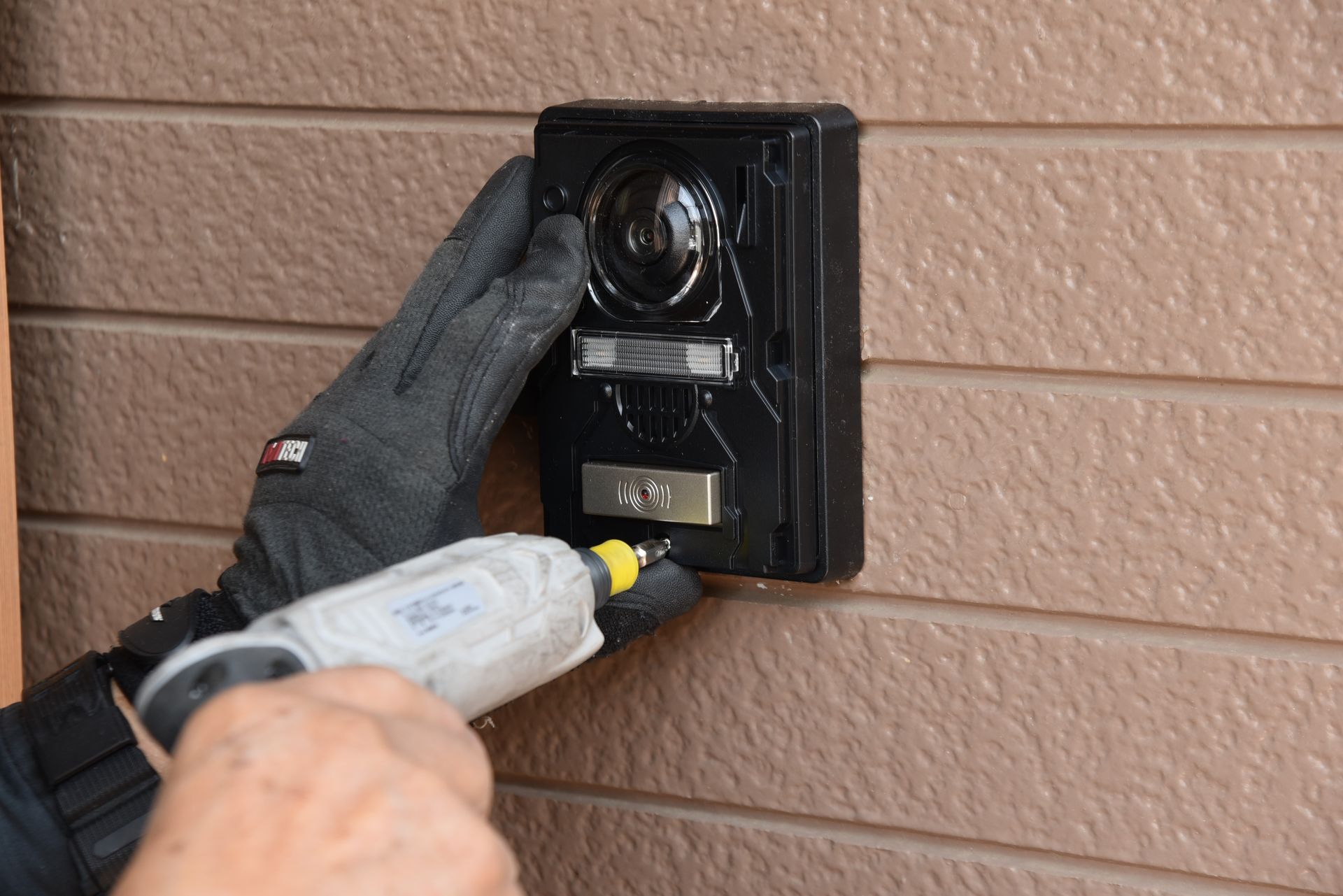 Person installing a black doorbell with a camera on a brown wall, using a power drill.