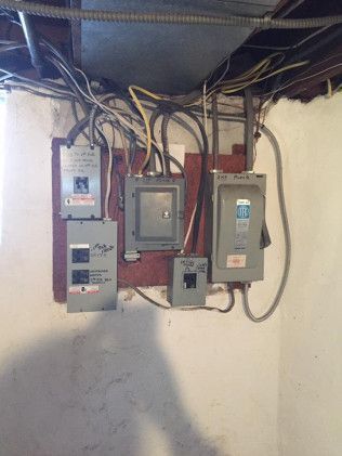 Electrical panel with various gray boxes and wires mounted on a wooden board against a white wall.