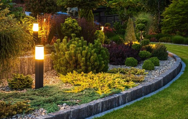 Illuminated garden bed with a variety of shrubs and ornamental grasses, edged with stone and illuminated by vertical lights.