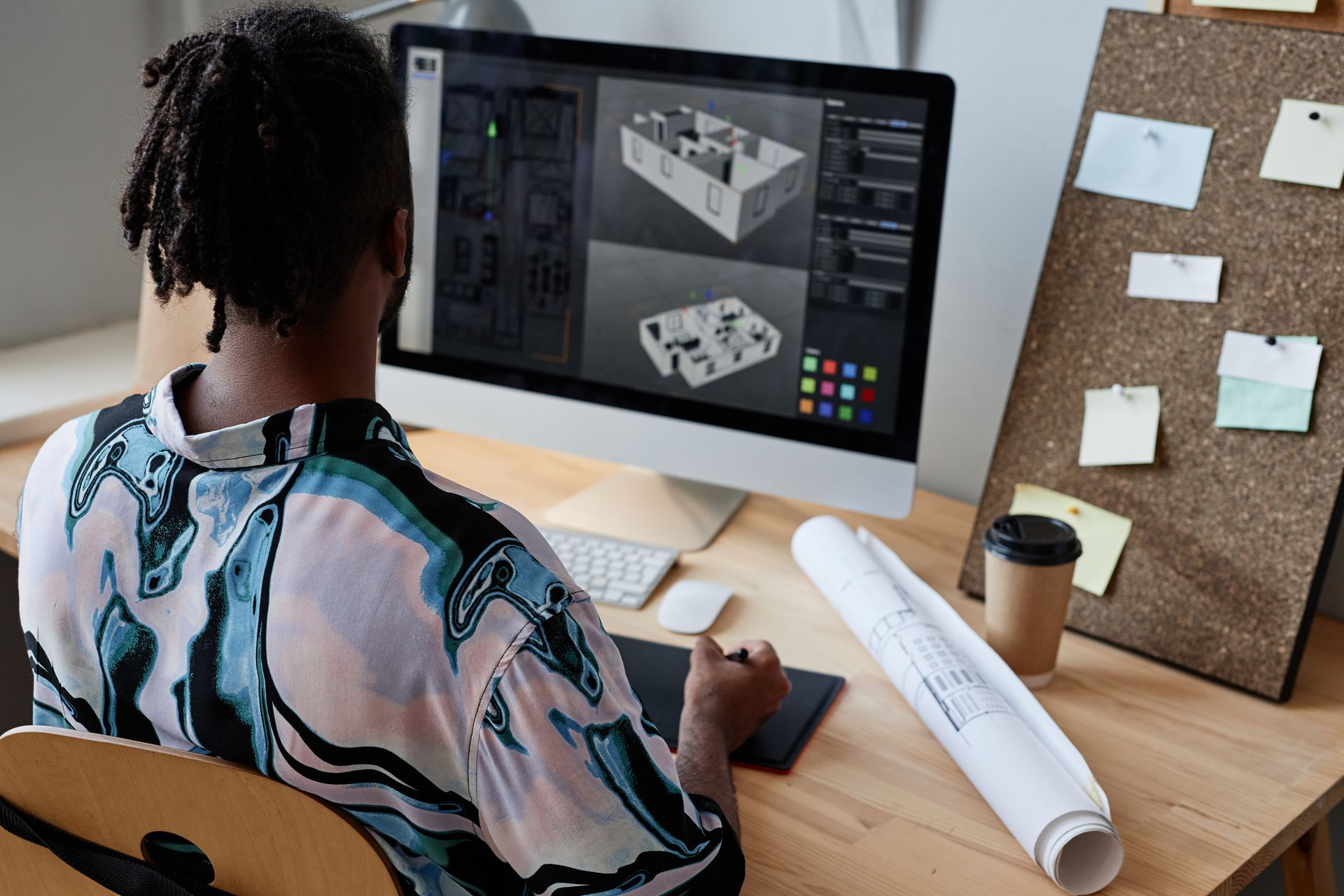 Person working on a computer showing a 3D architectural model, with a design tablet, blueprint, and bulletin board.
