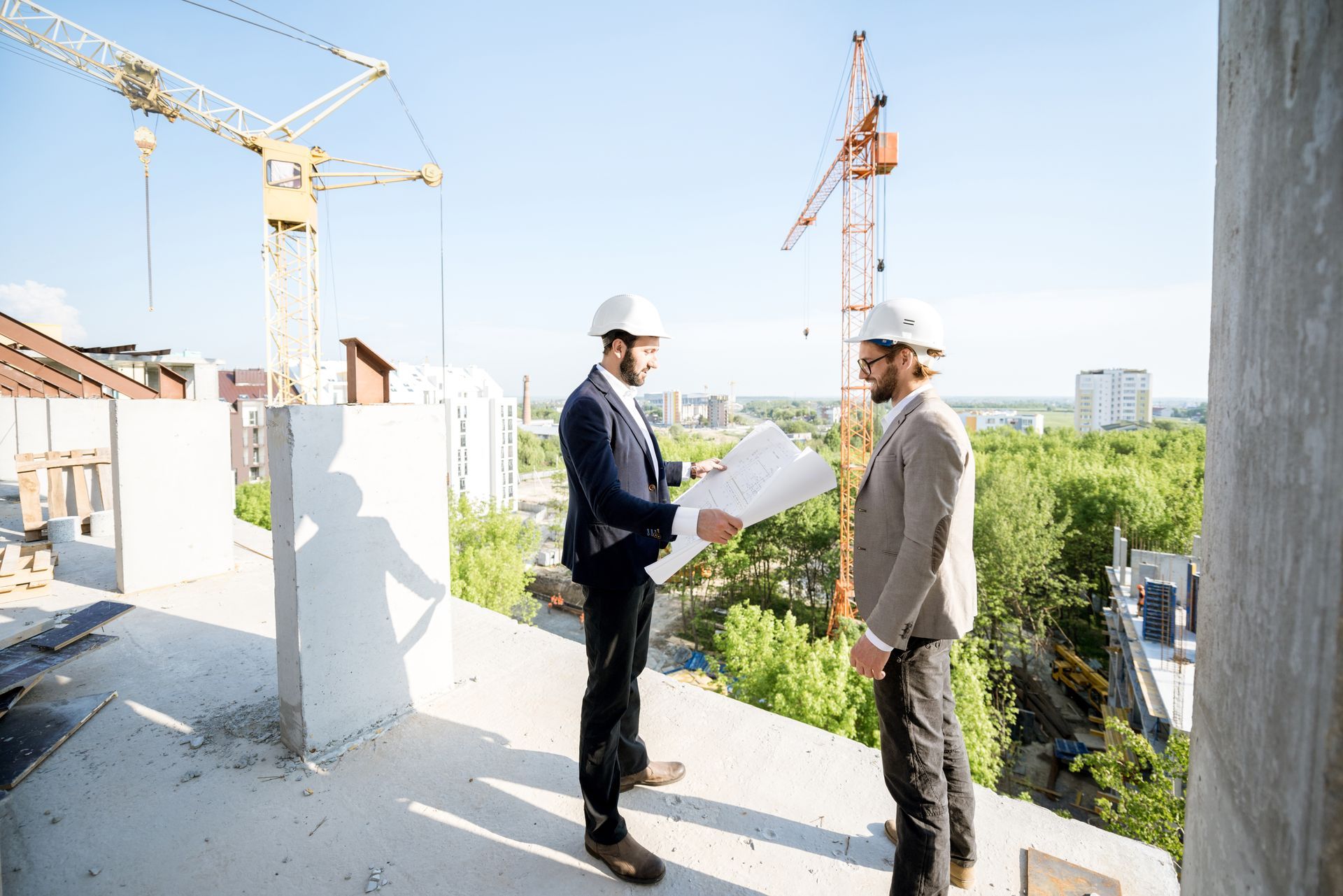 Two men in suits and hard hats on a construction site, reviewing blueprints.