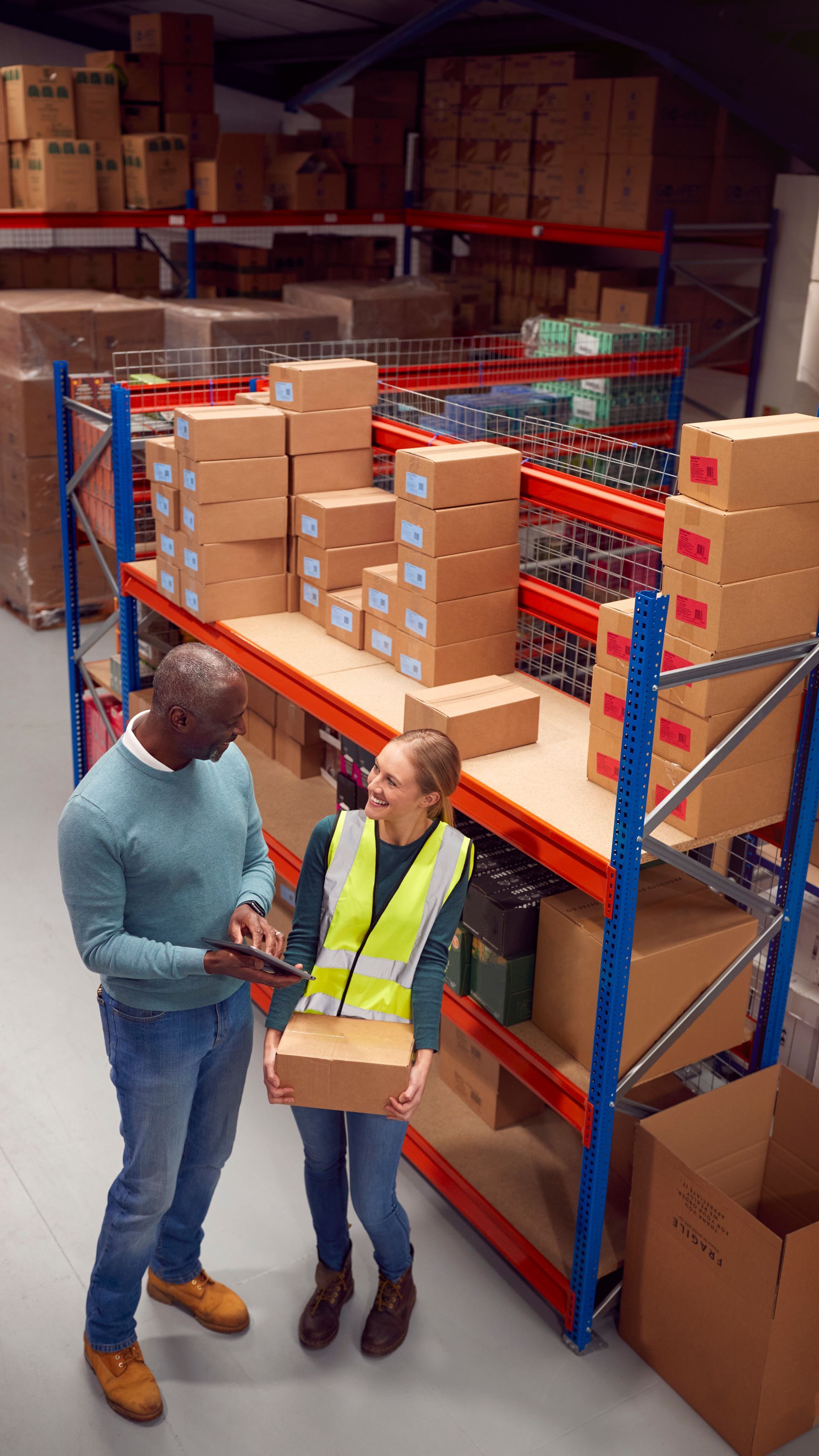 Two people in a warehouse: one holds a box, the other looks at a tablet. Shelves filled with boxes are in the background.