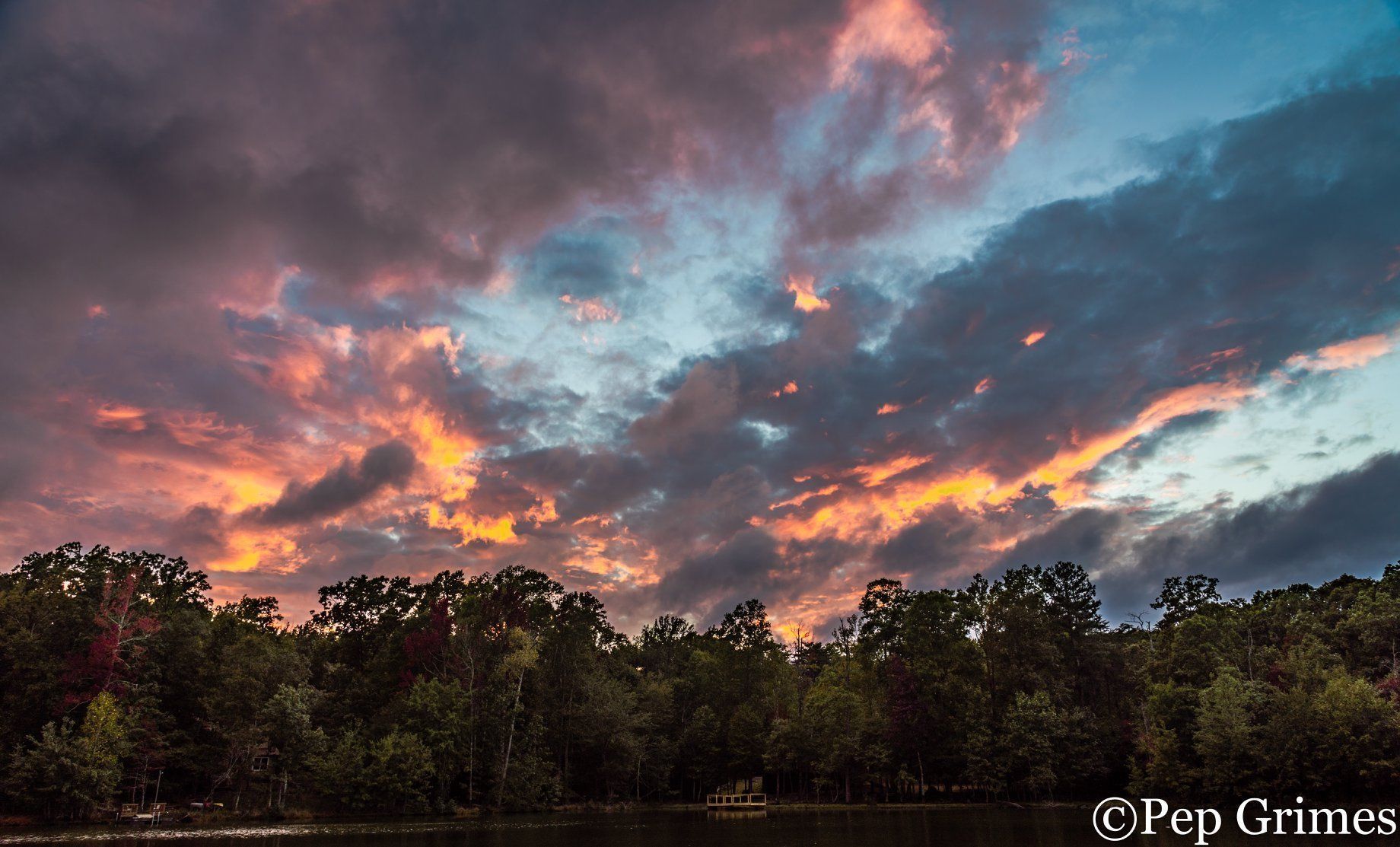 A sunset over a lake with trees in the foreground and a cloudy sky in the background.