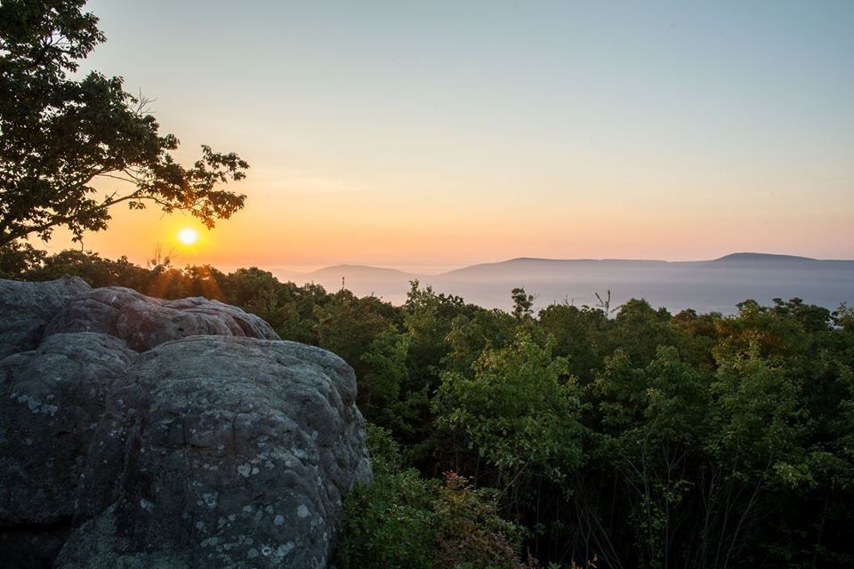 A sunset over a forest with a large rock in the foreground.