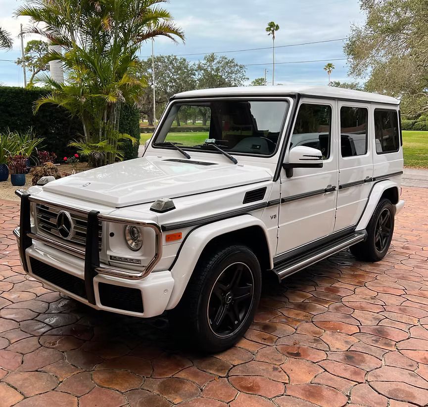 White Mercedes-Benz G-Wagon SUV parked on a paved driveway in front of a house. Black wheels, grille, and trim.
