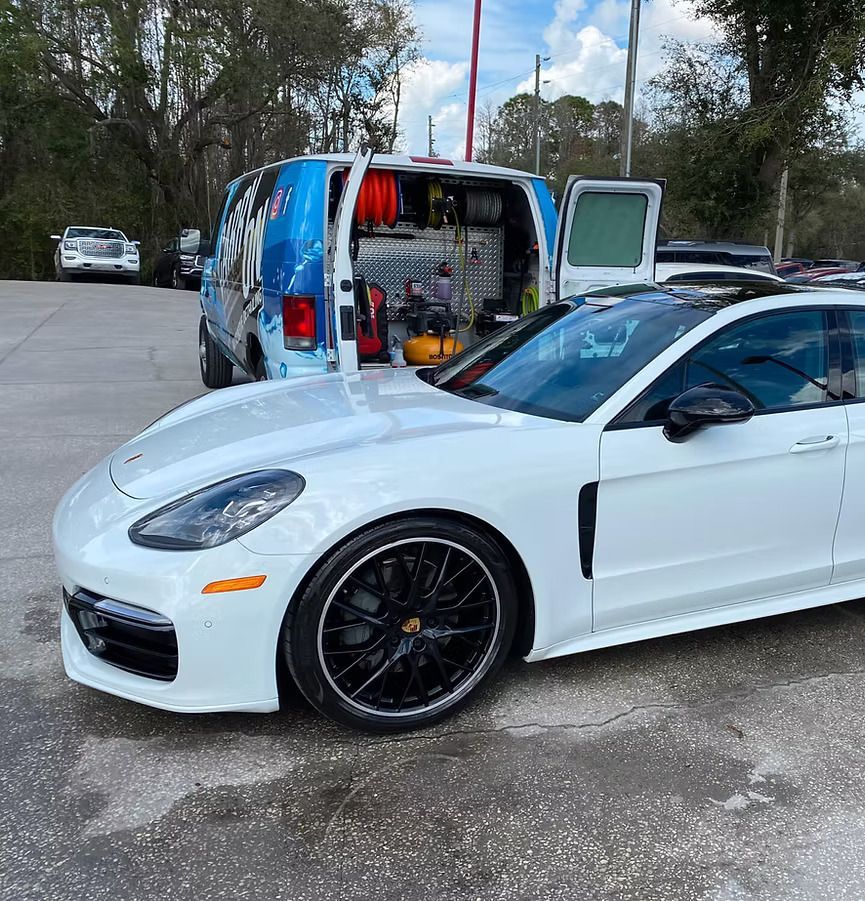 White Porsche Panamera next to a service van. The van's doors are open, revealing tools.