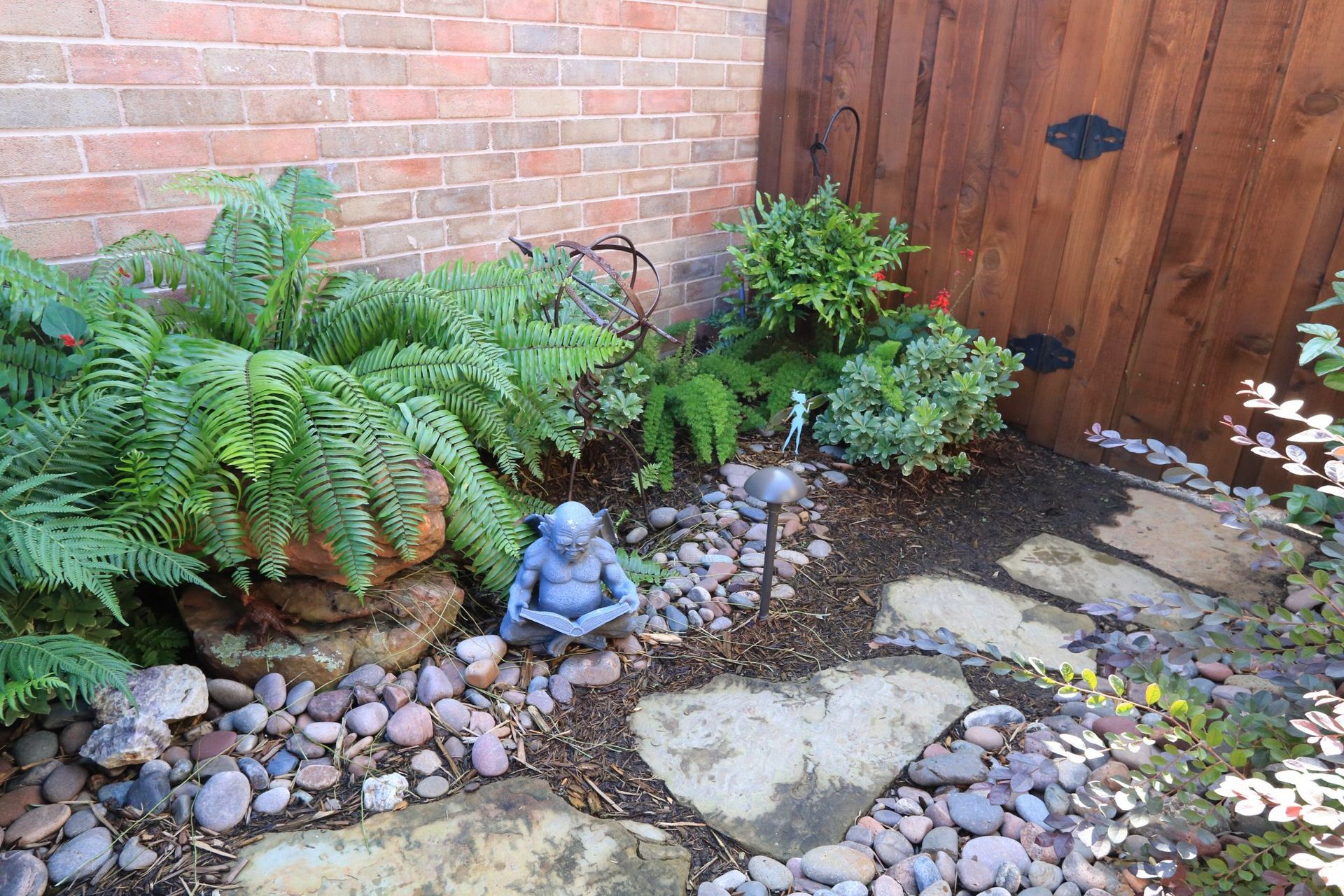Stone path leads past ferns, a meditating statue, and a wooden gate in a garden setting.