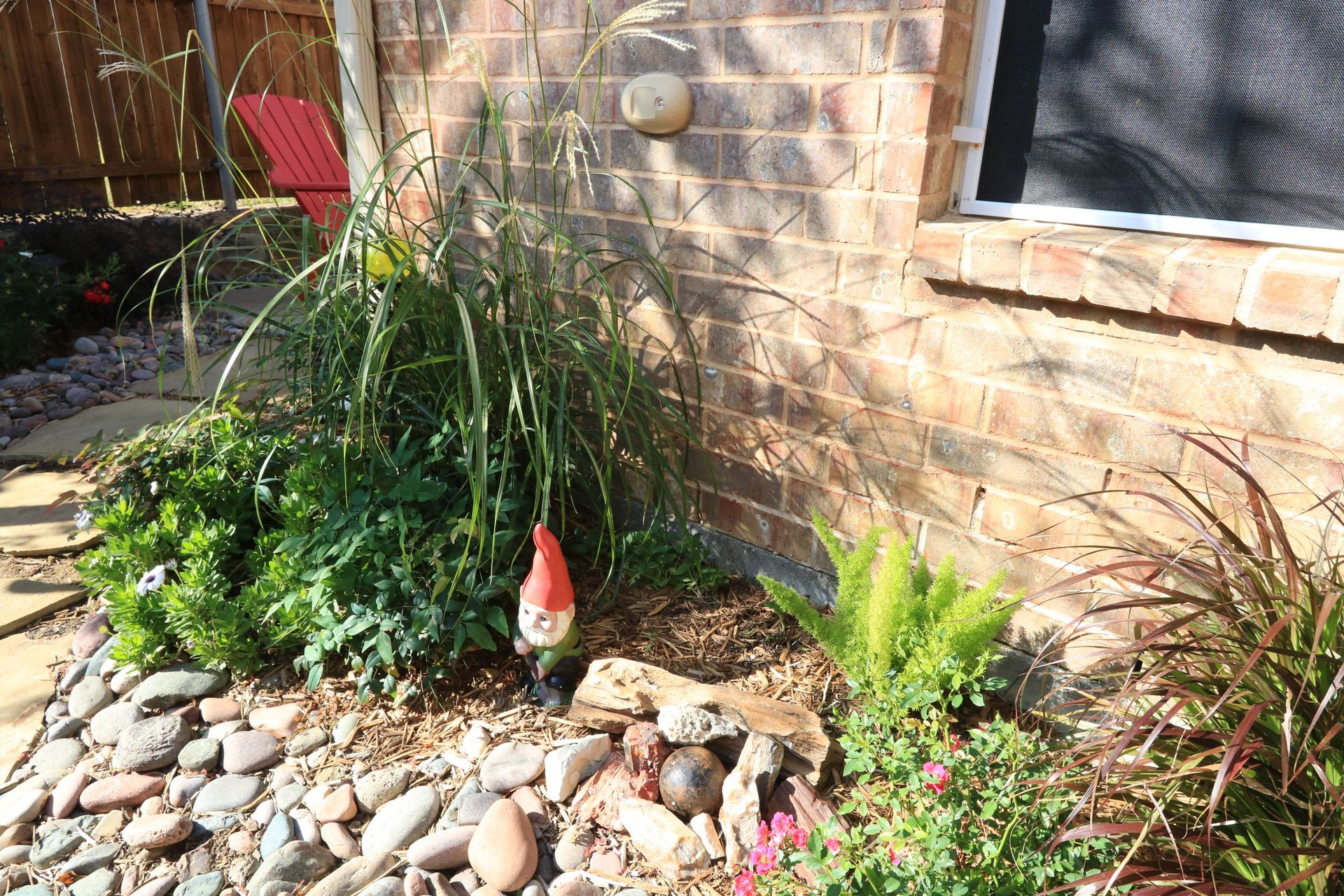 A garden bed with green plants and a gnome next to a brick wall, with a red chair in the background.