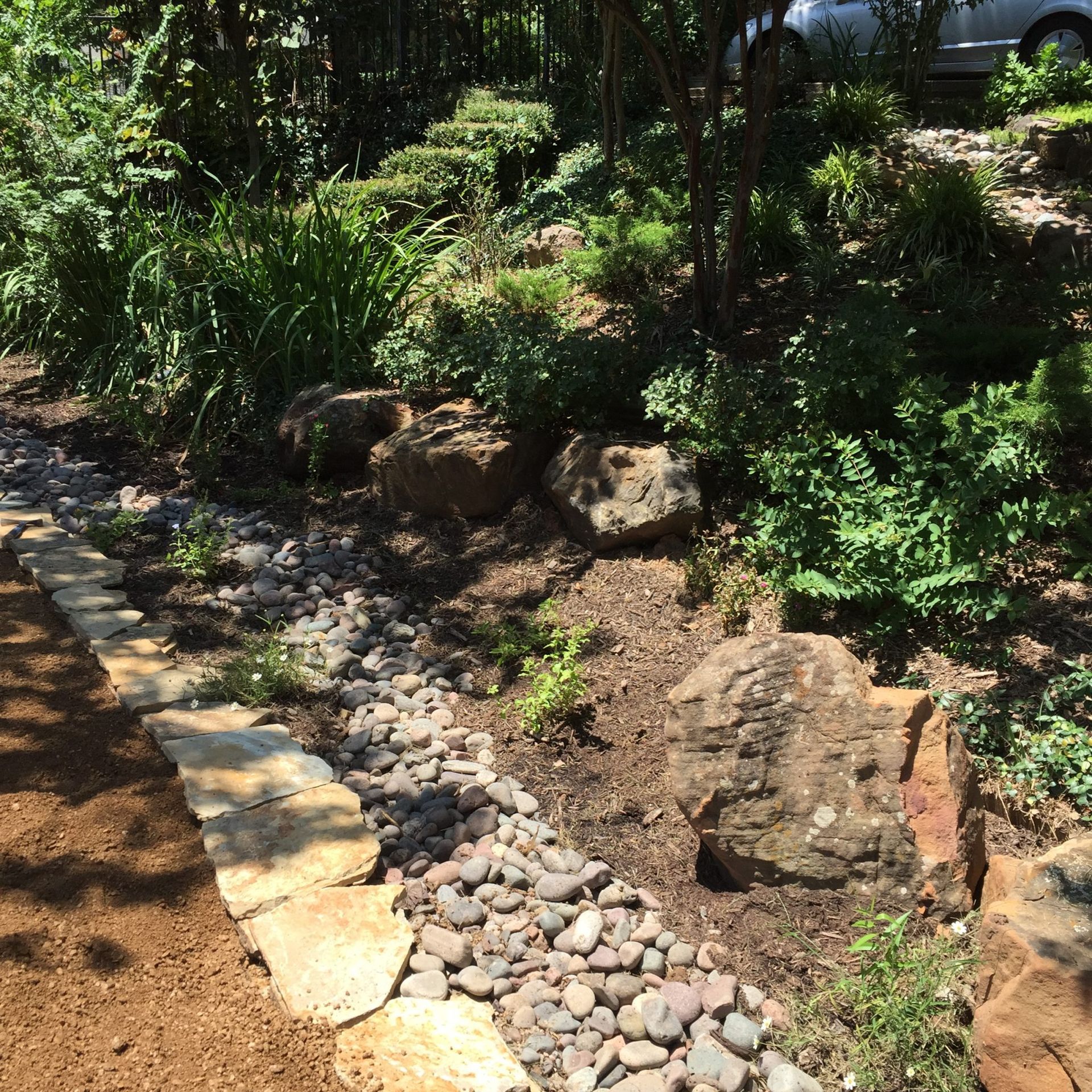 Stone-lined garden bed with rocks, plants, and a bordering path. Green vegetation and brown soil.