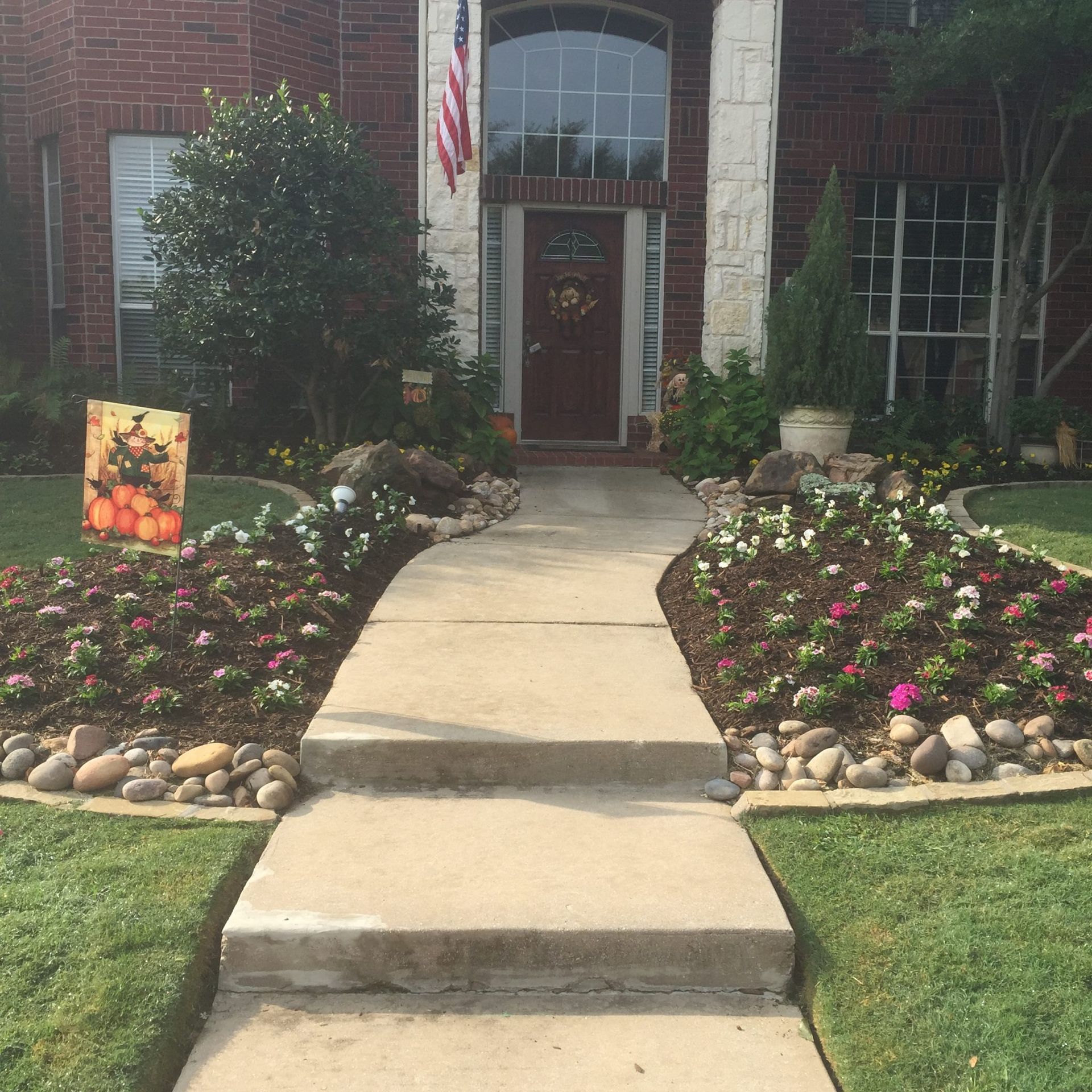 Concrete walkway to a house with flower beds, flag, and autumn decor.
