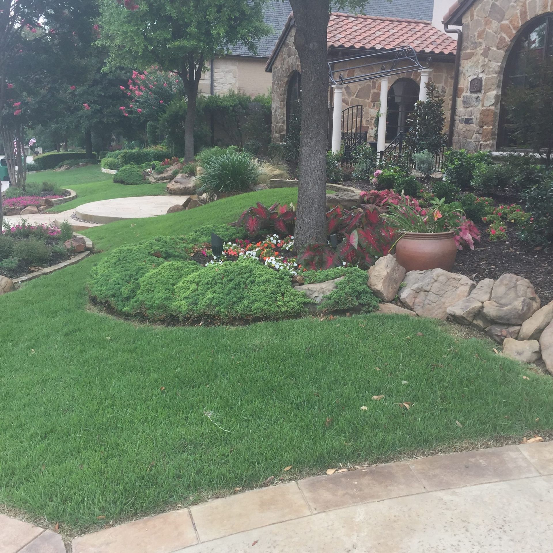 Well-manicured front yard with green lawn, trees, flowers, and a stone house.