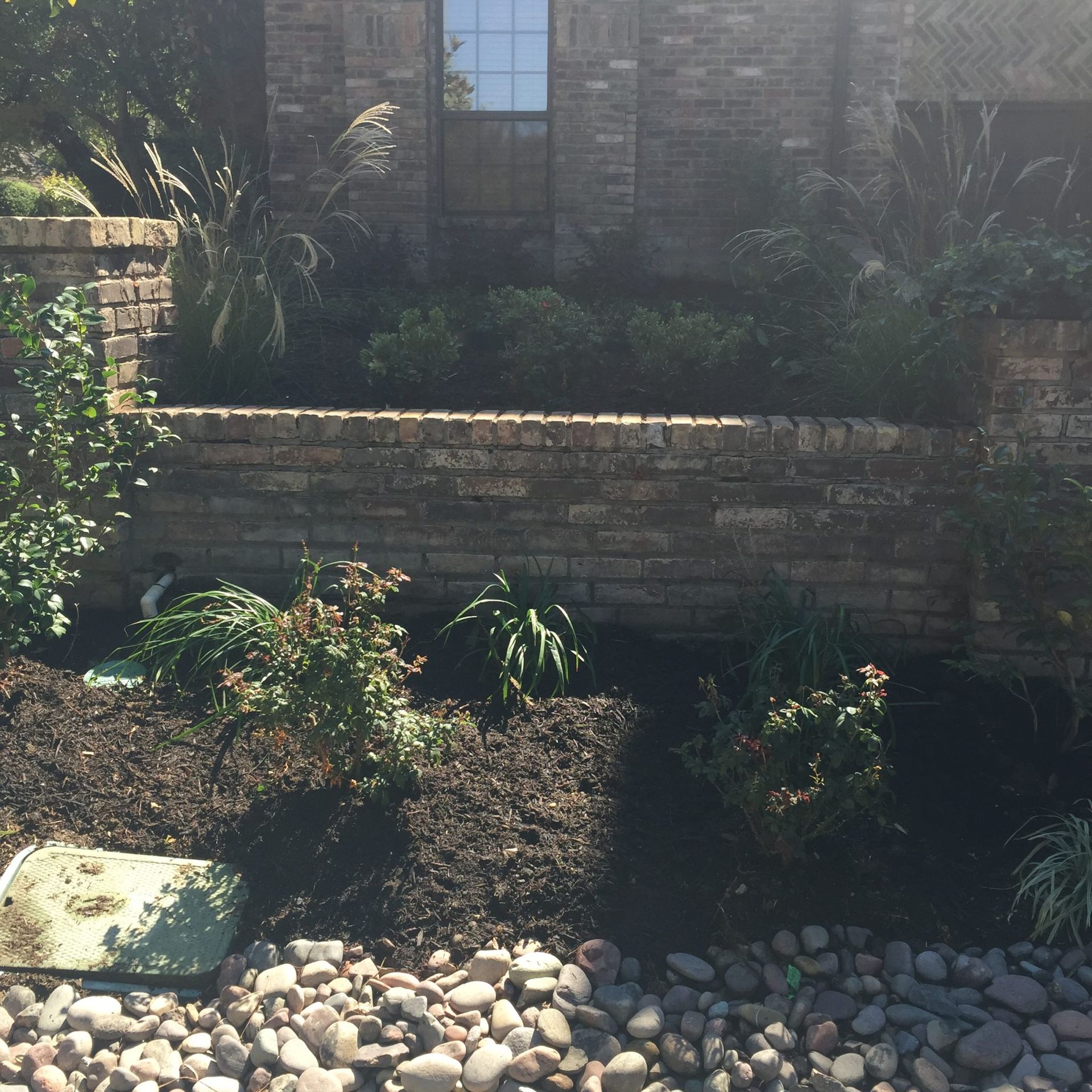 Brick wall with a landscaped bed; dark mulch, plants, river rocks.
