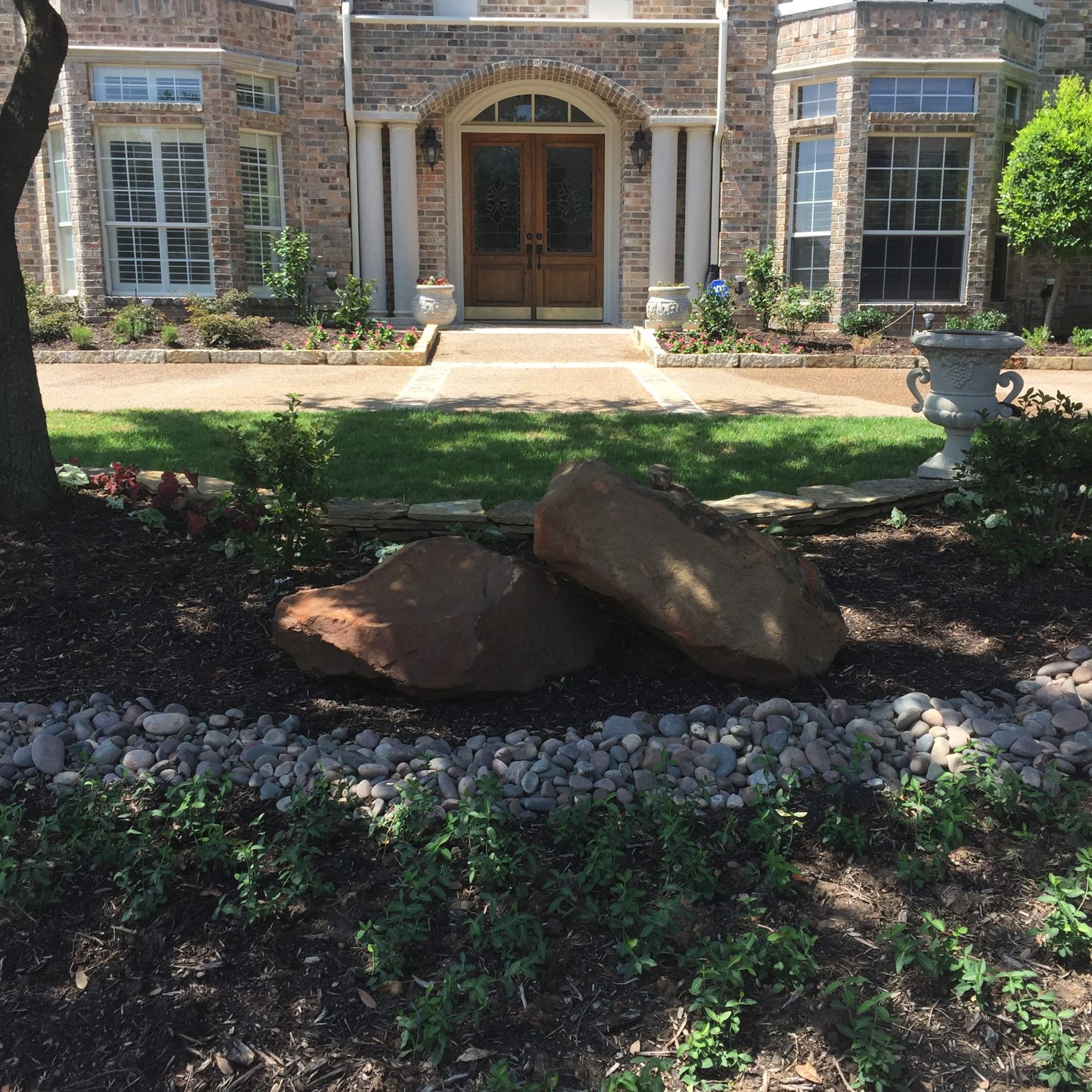 Two large brown rocks in a garden bed in front of a brick house with white columns and arched wooden doors.