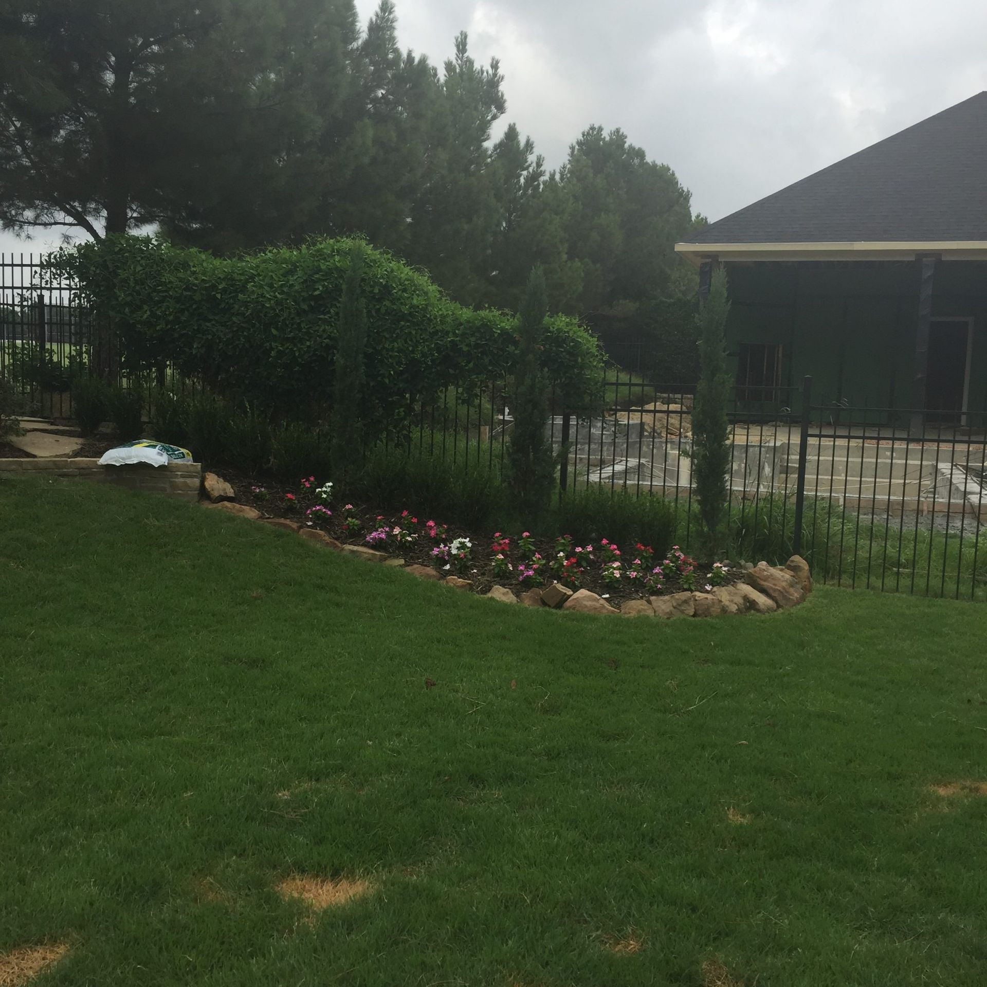 Green lawn with a flowerbed, bushes and trees, and a dark building and fence in the background.