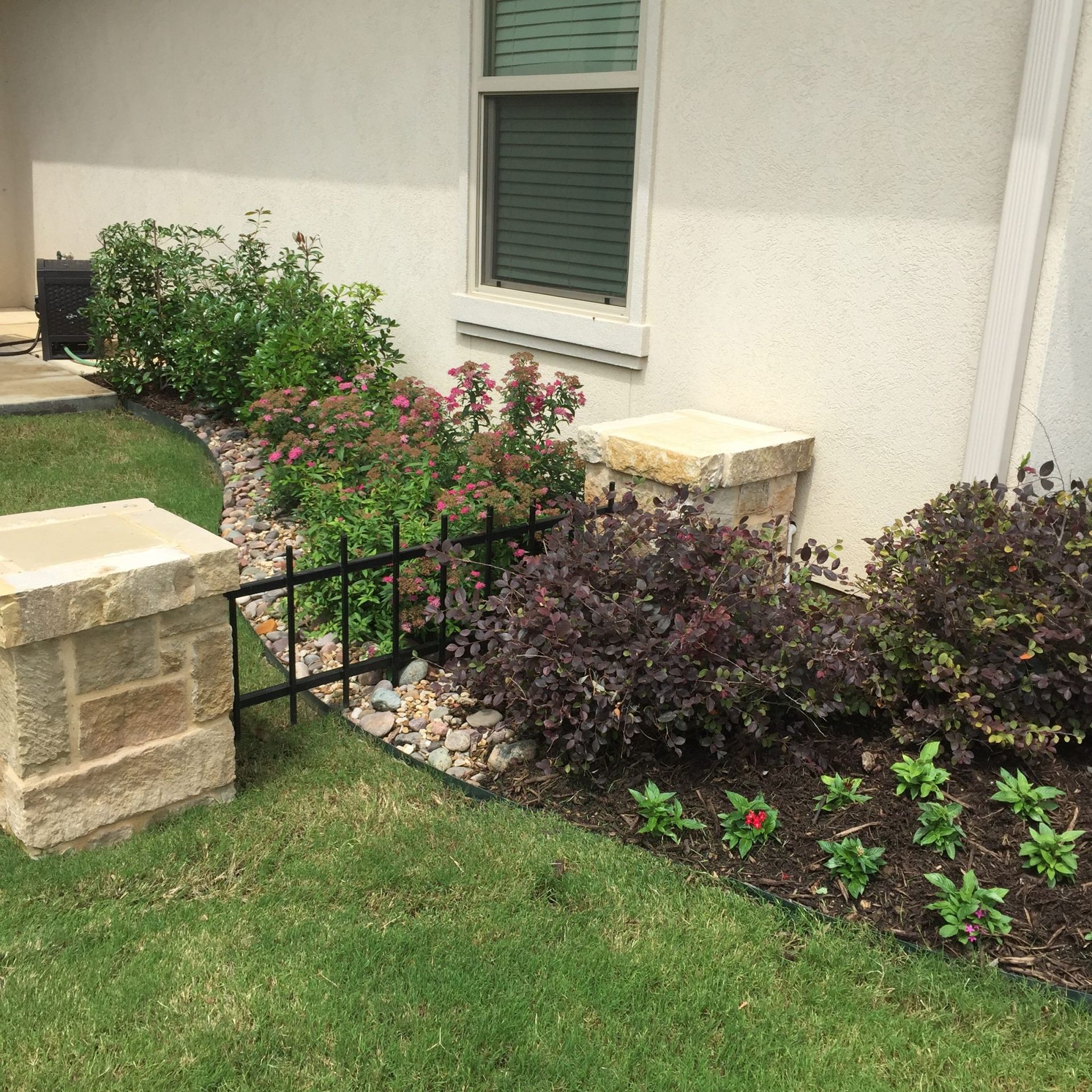 Garden bed with various plants, black fence, stone columns, and a window on a beige building.
