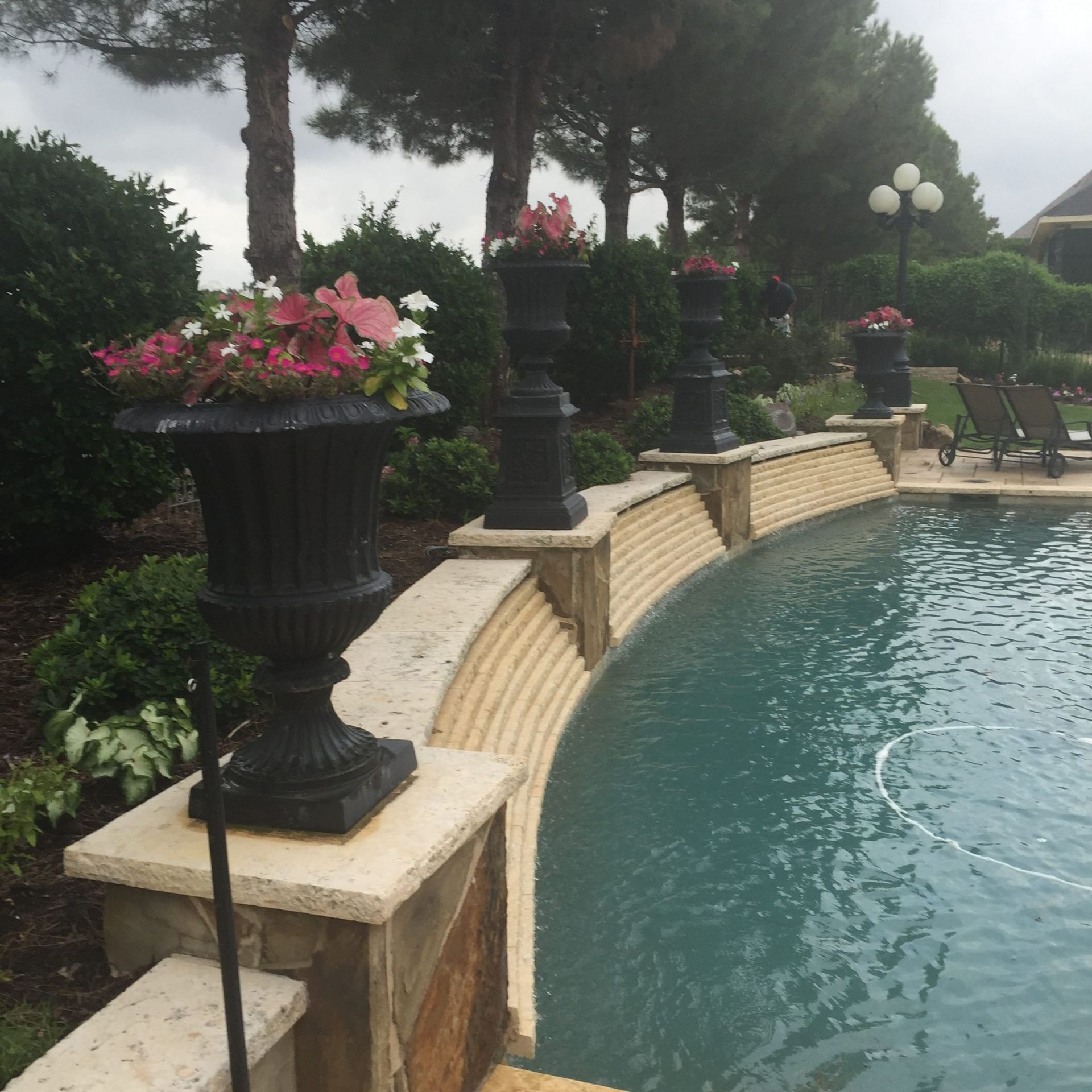 Poolside with urns of flowers along a curved wall, trees and a cloudy sky in the background.