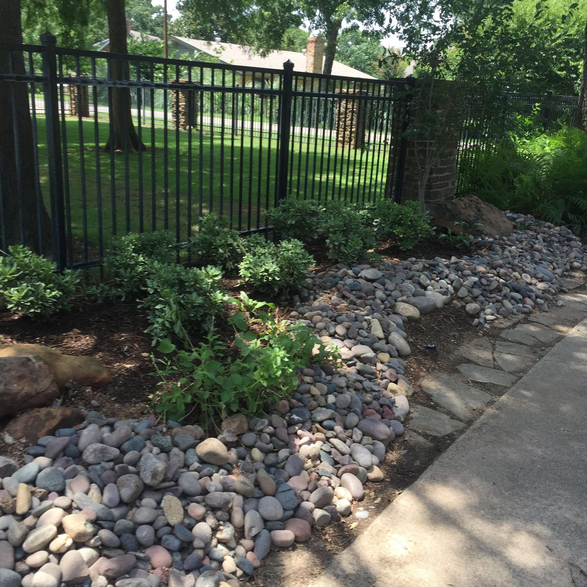 Black fence, rock border, bushes, and plants near a sidewalk.