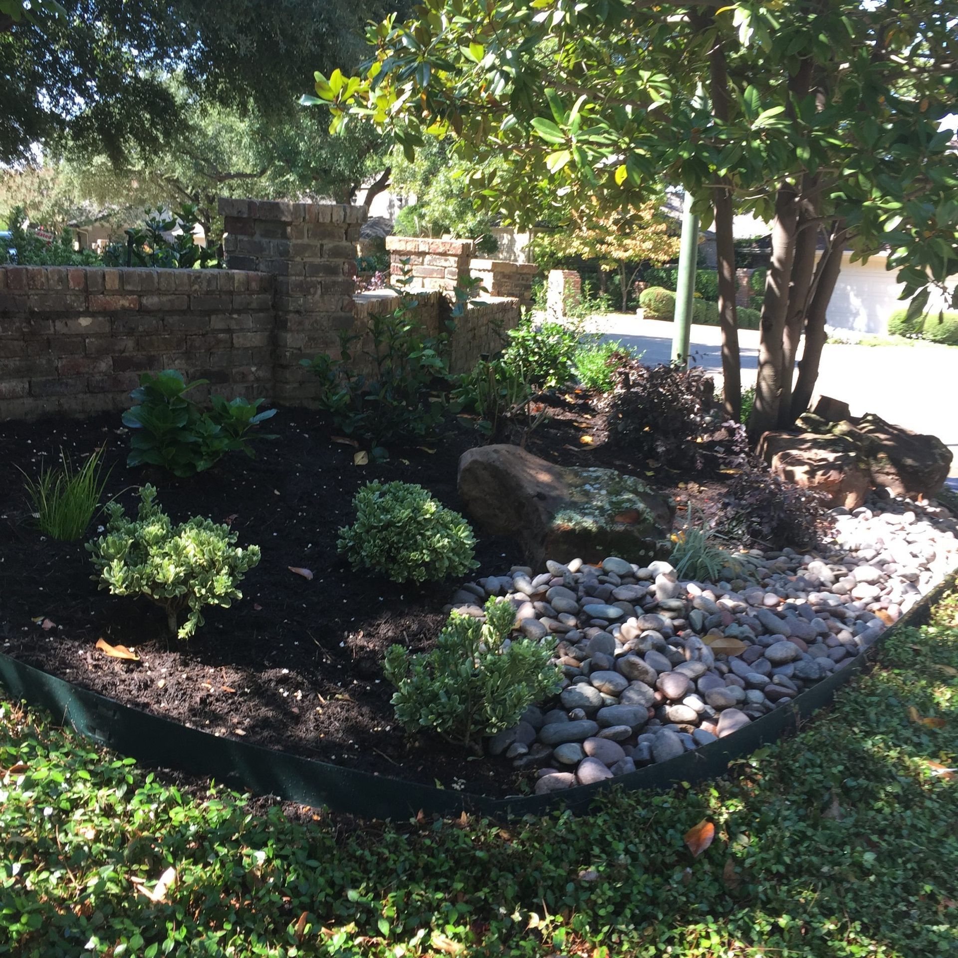 Landscaped garden bed with plants, rocks, dark mulch, and a brick wall.