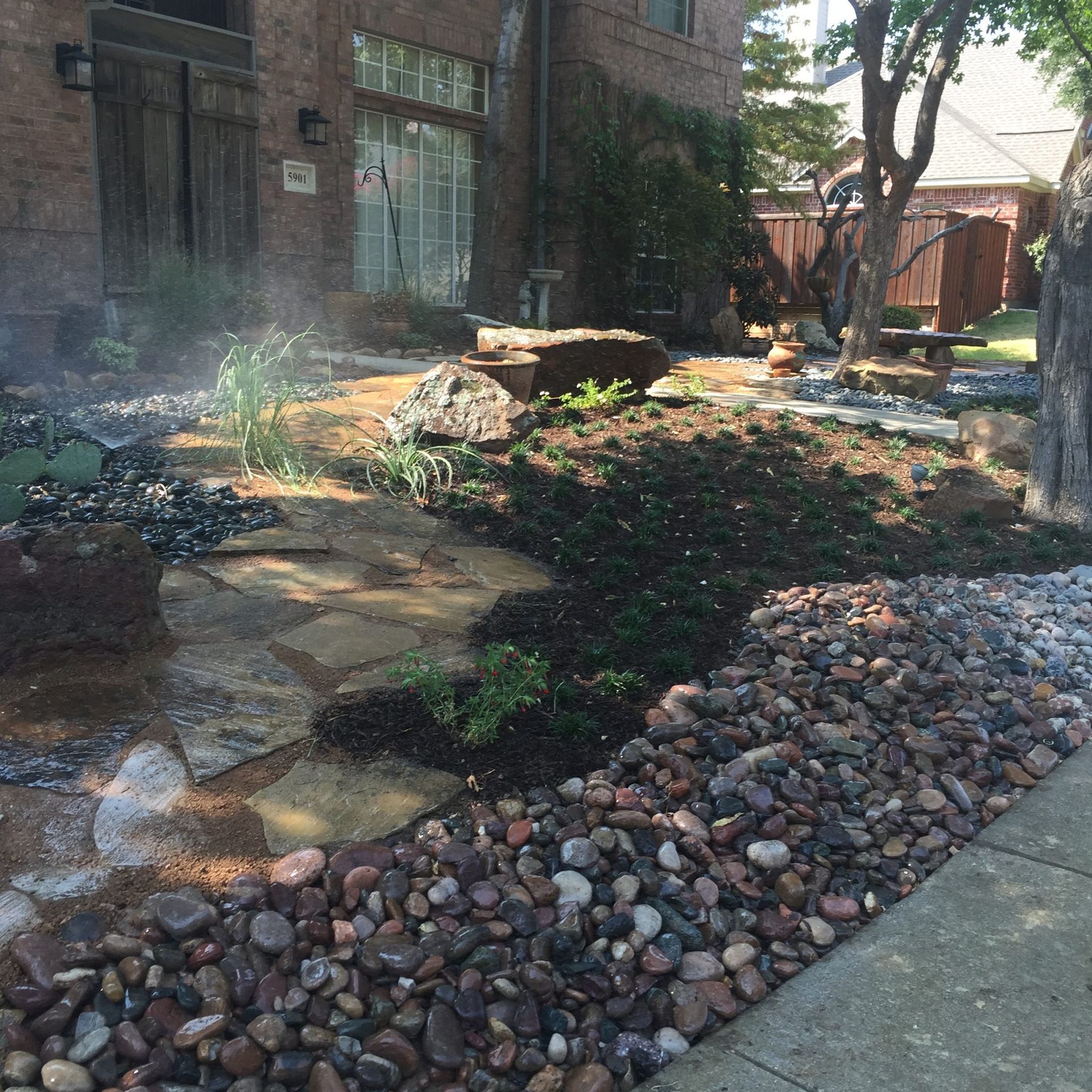 Stone walkway and landscaping next to brick building and sidewalk, with rocks, plants, and a water feature.