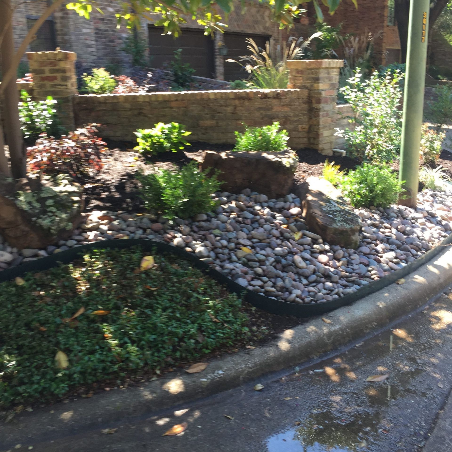 Landscaped garden bed with rocks, plants, and a brick wall along a street.