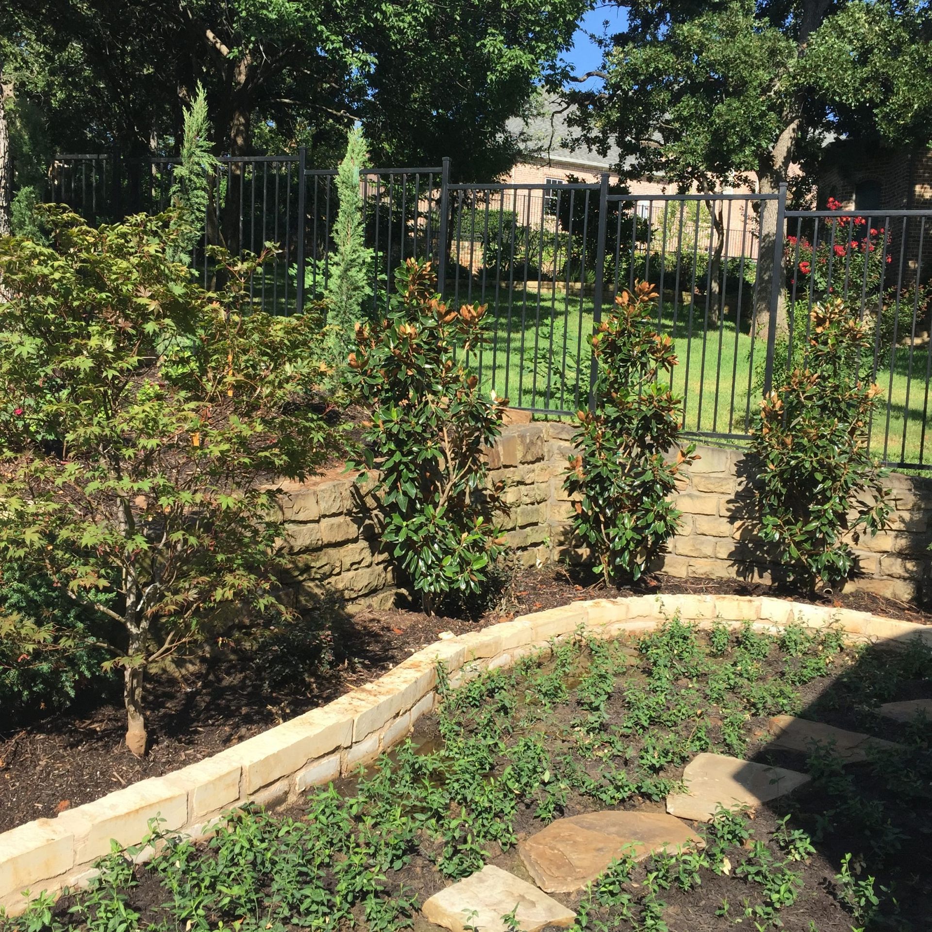 A landscaped backyard with a stone wall, shrubs, and a black metal fence.