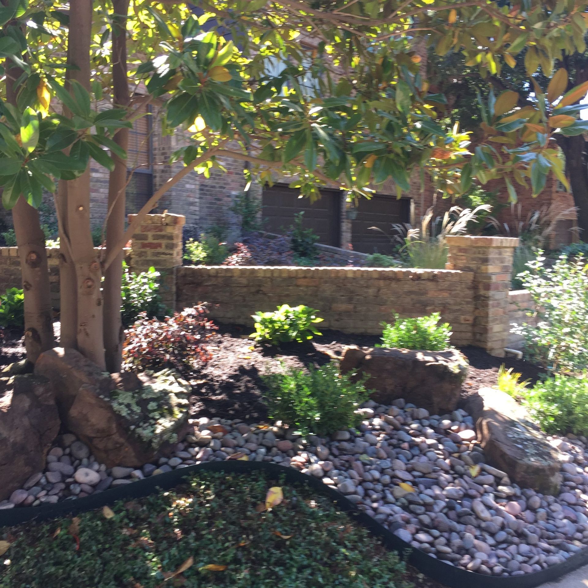 Landscaped front yard with brick wall, large rocks, and various greenery under trees.