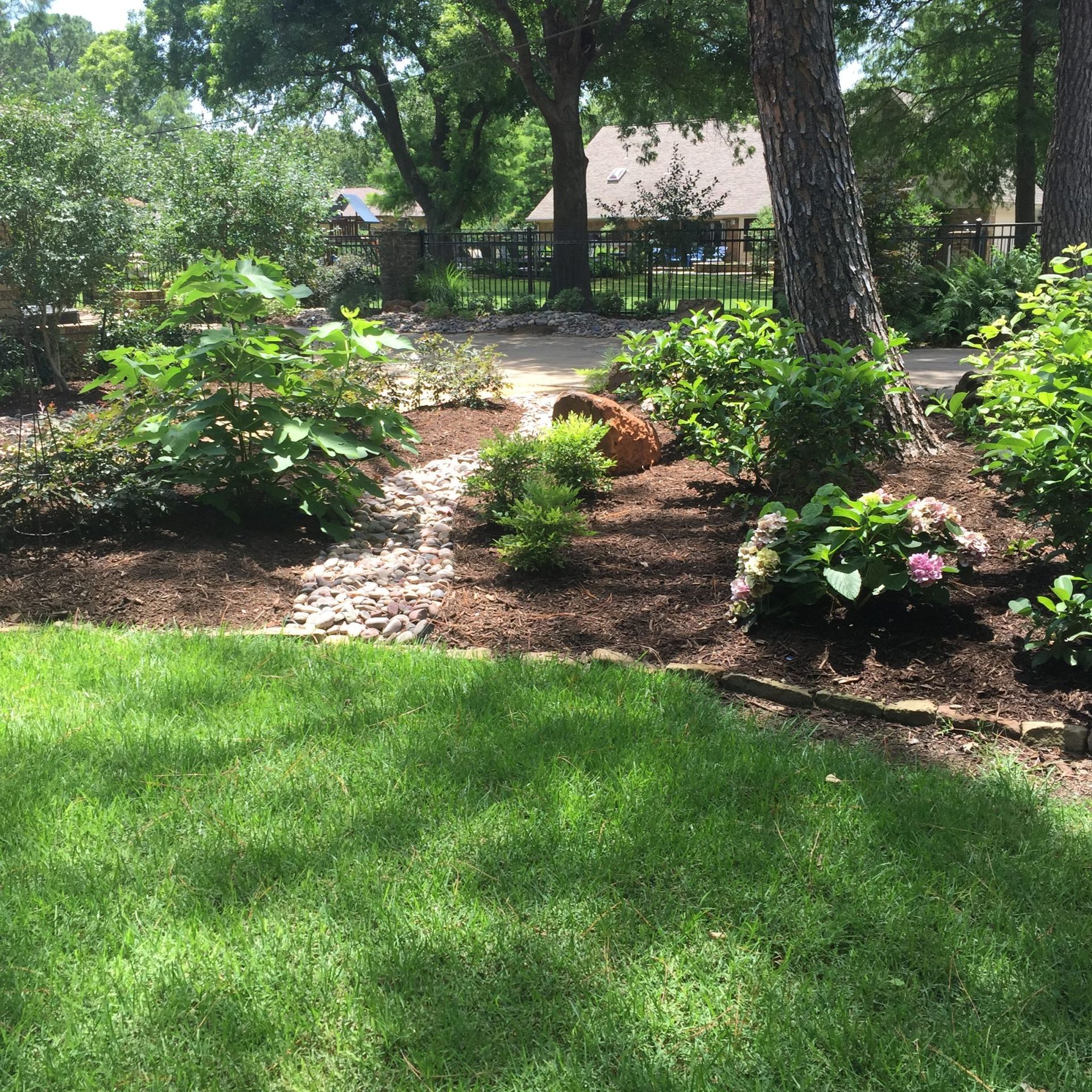 Lush green yard with a stone-lined dry creek bed, trees, and flowering bushes.