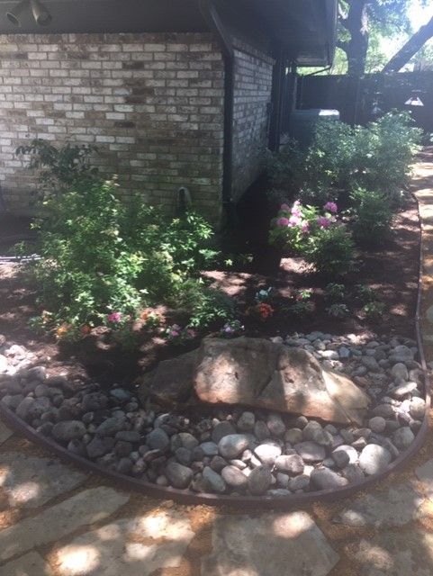 Stone landscaping with mulch, shrubs, and a large rock border a building's brick exterior.