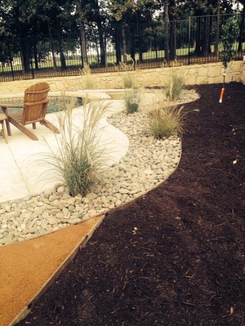 Landscape with a concrete patio, decorative rocks, mulch, and ornamental grasses by a pool.