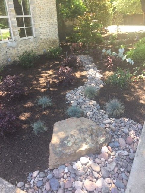 Rock-lined dry creek bed with landscaping and plants beside a stone house.