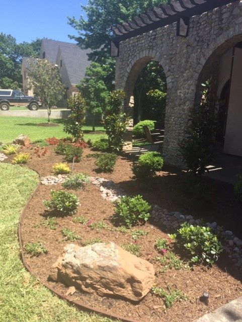 Landscaped garden bed with brown mulch, rocks, and green bushes near stone archway.