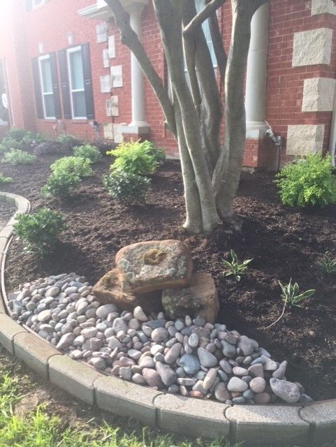 Landscaped front yard with stone edging, mulch, river rocks, bushes, and a tree in front of a brick house.