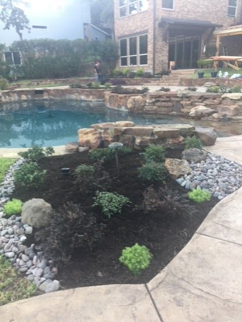 A landscaped pool area with rocks, plants, dark mulch, and a large brick home in the background.