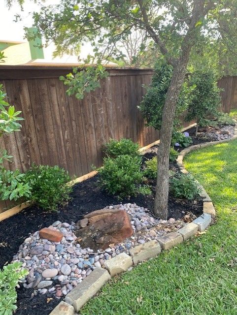 Lush backyard garden bed with brown mulch, rocks, green bushes, and a tree next to a wooden fence.