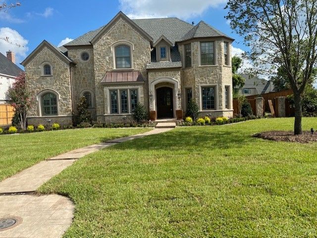 Two-story stone house with a green lawn, sidewalk, and a few trees under a blue sky.