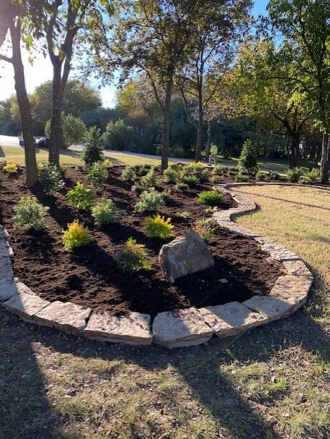 Stone-edged garden bed with mulch and various green plants, a large rock, under trees; outdoors.