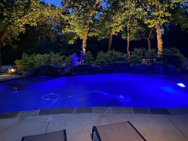 Nighttime pool with blue light, rock waterfall, and trees illuminated with spotlights.