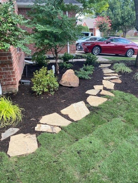 Stone path leads through a landscaped yard with plants and a large rock, next to a brick house.