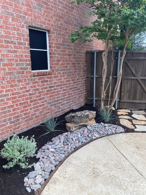 Brick wall with landscaping including rocks, plants, and a small tree next to a patio.