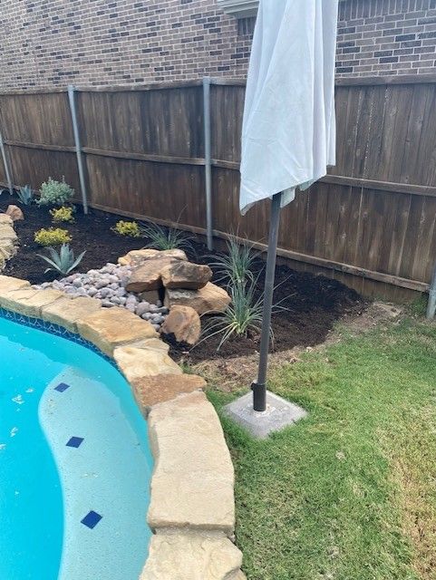 Poolside landscaping with plants, rocks, dark mulch, umbrella near a brown fence.