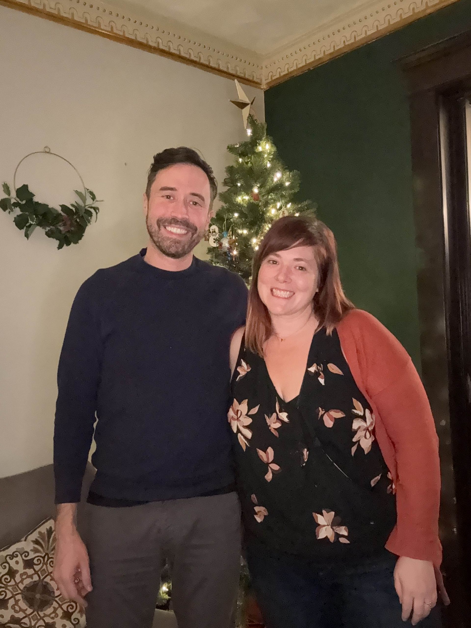 Two people—a white man and a white woman—smile in front of a Christmas 